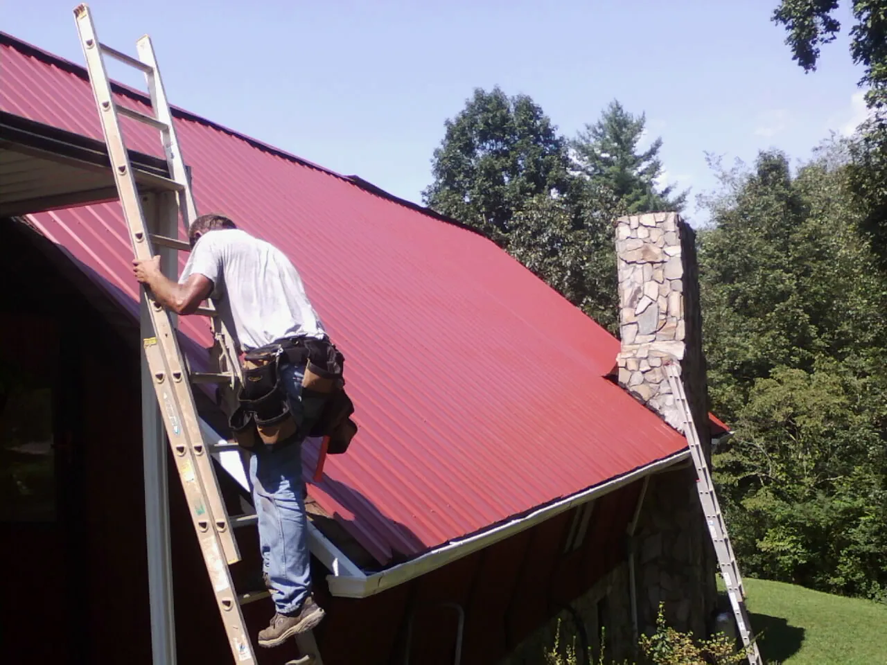 Roofer on a ladder beside a red metal roof and stone chimney — Soffit & Fascia Repair by Roofing Brothers in Brunswick